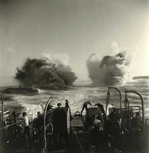 Black and white photograph. The stern of a large ship, men stand with their backs to the camera. In the distance, plumes of water shoot aggressively into the air as depth charges are exploded.